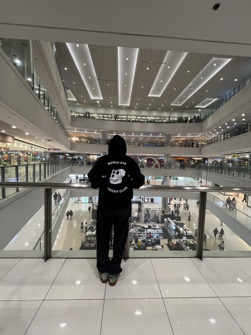 The image shows a person standing alone inside an empty mall, wearing a hoodie with a skull design on it. The individual appears bored or disinterested as they stand near a glass railing overlooking the interior of the mall. The surrounding area consists of multiple stores and people walking around, but the main focus is on this person in their hoodie.

The post suggests that the person might be interested in joining an exclusive club called "Bored Ape Yacht Club". This club offers memberships to various yacht clubs worldwide, providing access to exclusive yachts and experiences for its members. The image captures a moment of contemplation or boredom as the individual ponders whether they should join this club or not.