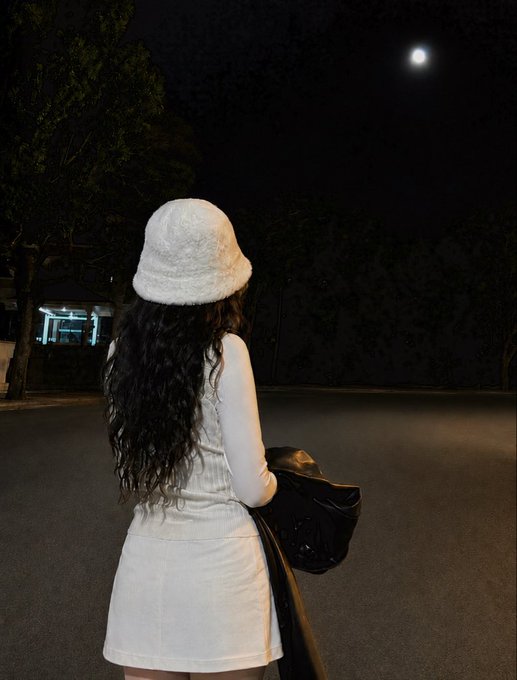 A person wearing a white bucket hat, white long-sleeve top, and white skirt stands facing away from the camera, looking at the moon in the night sky while holding a black jacket.