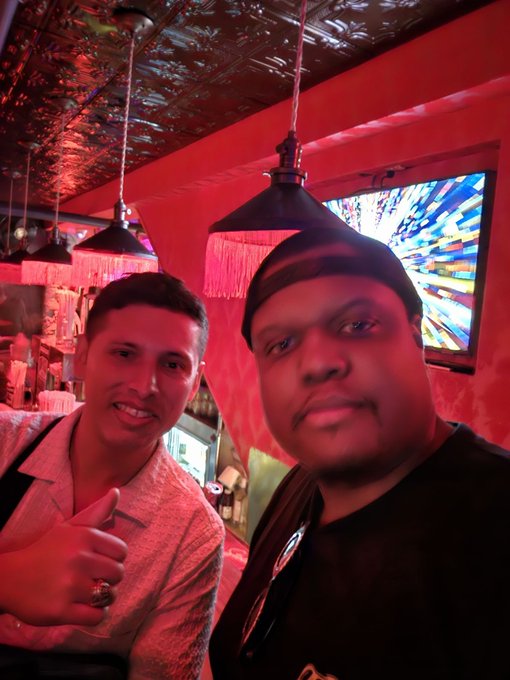 A selfie of two men inside a red-lit venue with decorative hanging lamps and a stained-glass window in the background.