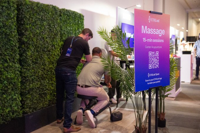 The image shows a man sitting on a chair, receiving a 15-minute chair massage from another person standing behind him. The scene takes place inside an office setting with a green hedge wall as a backdrop. There are two people present in the room - one seated and one standing near the chair massager. A sign is displayed nearby that reads "Massage" and "15 min".