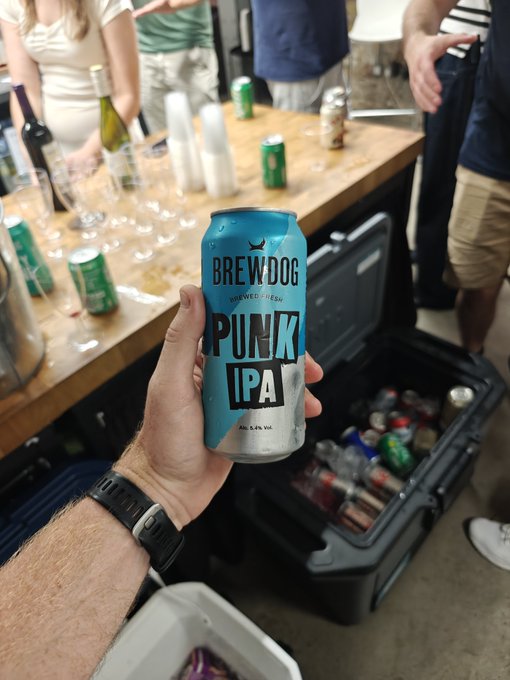A hand holds a blue can of BrewDog 'PUNK IPA' in the foreground, with a wooden table, other drinks, and people gathered behind it.