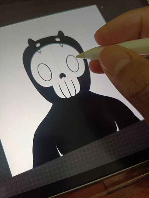 A hand using an Apple Pencil to digitally draw a black bear-like character with a skull face on a tablet screen.