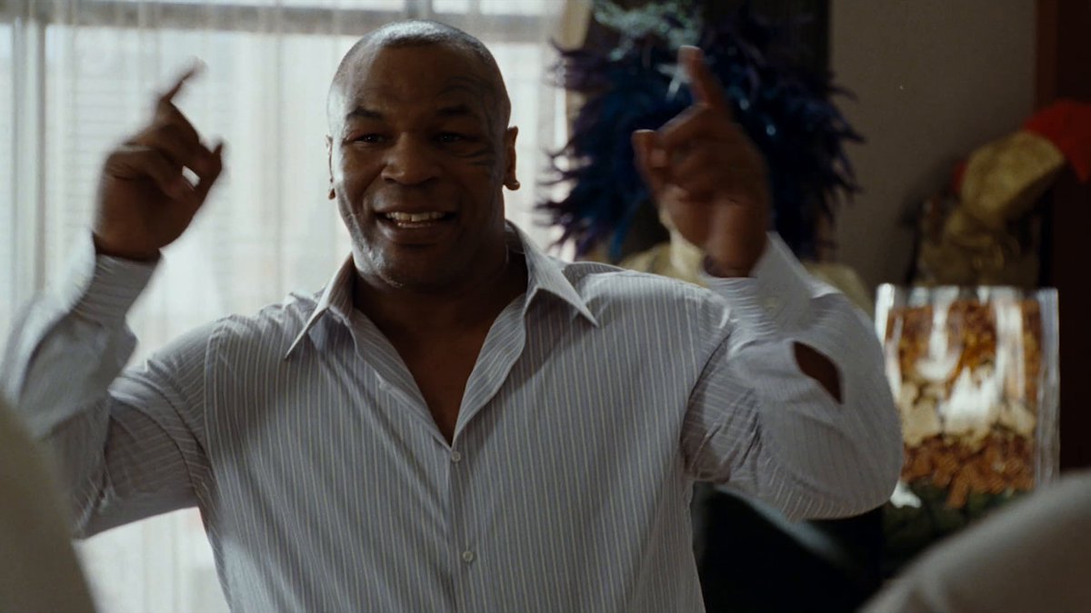 A scene from a movie featuring Mike Tyson in a striped button-up shirt, smiling and raising both hands with his middle fingers extended.