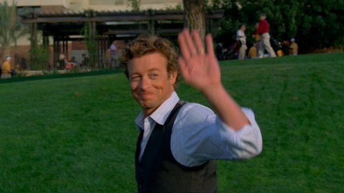 A man with curly blonde hair, wearing a light blue shirt and dark vest, smiles and waves at the camera in an outdoor grassy setting with people in the background.