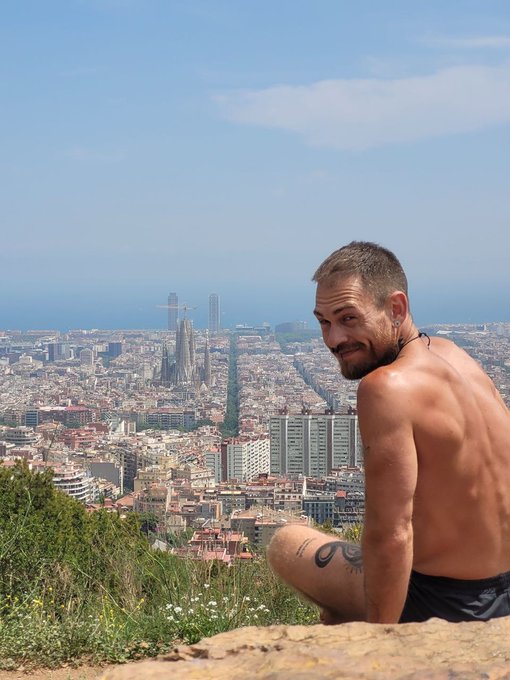 A shirtless man with a beard sitting on a hilltop overlooking Barcelona, with the Sagrada Família visible in the cityscape below.