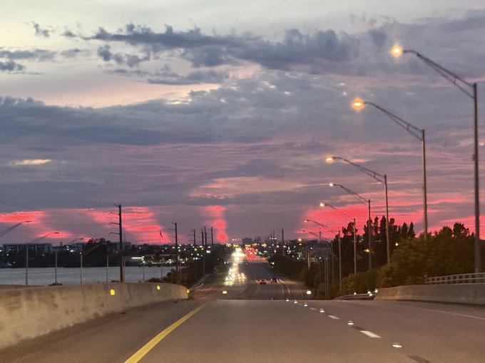A highway stretches toward the horizon under a vibrant pink and purple sunset sky, with streetlights lining the road and a body of water visible to the left.
