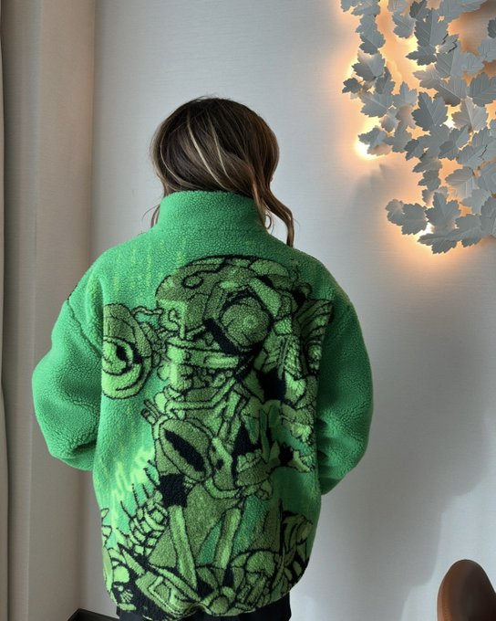 A person photographed from behind wearing a bright green fleece jacket with a large black embroidered graphic design, standing near a decorative leaf wall light.