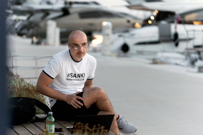 A bald man with glasses wearing a white t-shirt with the text '#SANKI' and 'SANKIHAJA.COM' sits at a marina with a laptop and a water bottle, with several yachts visible in the background.