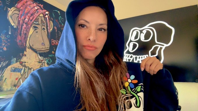 A woman wearing a blue hoodie poses in front of two artworks, including a painting of a Bored Ape Yacht Club character on the left and a neon skull sign on the right, connecting to the @BoredApeYC reference in her post about authentic roots culture.