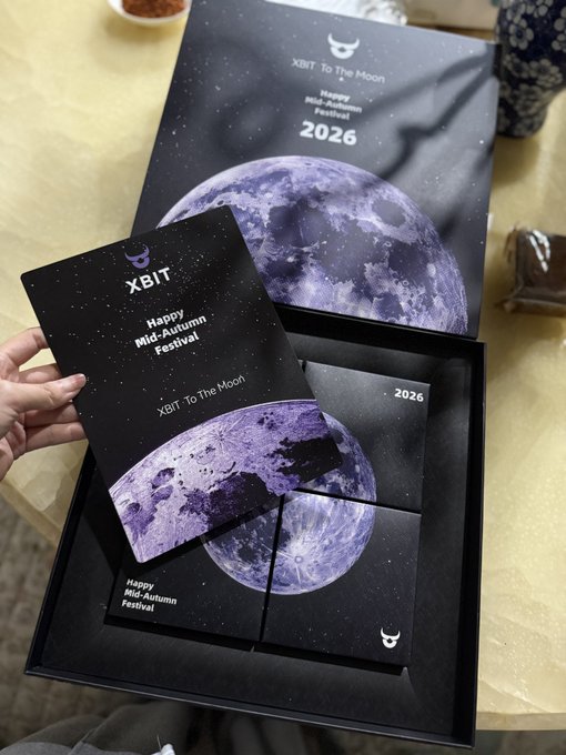 A hand holds a dark card in front of an open gift box set, featuring a moon design and the text 'XBIT - To The Moon' and 'Happy Mid-Autumn Festival 2026'.