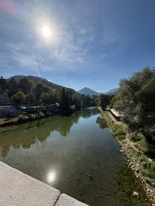 A serene river reflects the bright sun beneath a clear blue sky, framed by lush green mountains and trees that evoke the calm and tranquility essential for protecting one's mental health. The peaceful waterside view offers a natural reminder to slow down and reconnect with nature as part of self-care.