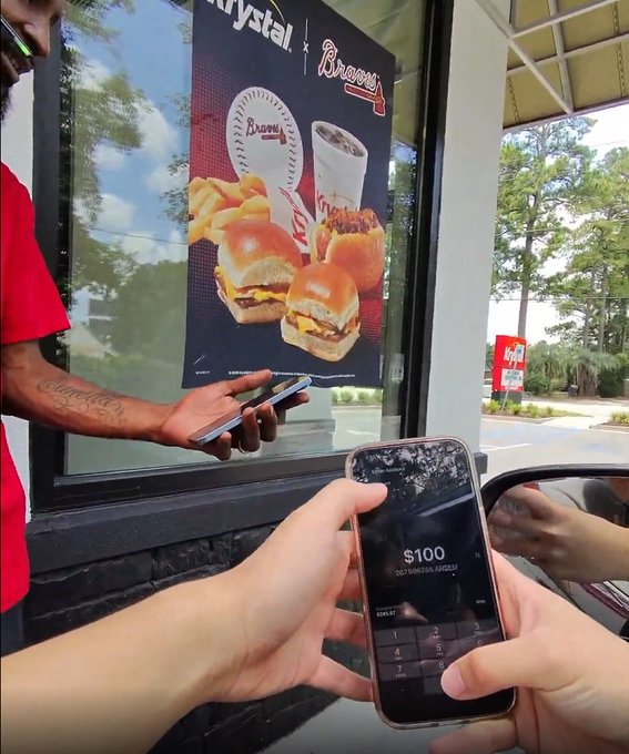 A person holds a smartphone displaying a $100 airdrop from "00F30031493232" to a Crystal/Atlanta Braves drive-thru employee, illustrating the creator's real-life crypto onboarding stunt for the $ANSEM token.