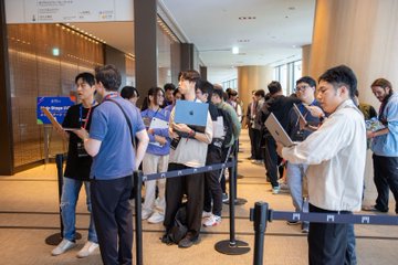 A group of people, many holding laptops and tablets, standing in a queue area divided by blue stanchion belts in what appears to be a tech conference or event venue.