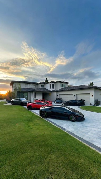 A modern two-story house at sunset with multiple luxury cars parked in the driveway, including a black Lamborghini and a red sports car, set against a dramatic sky.