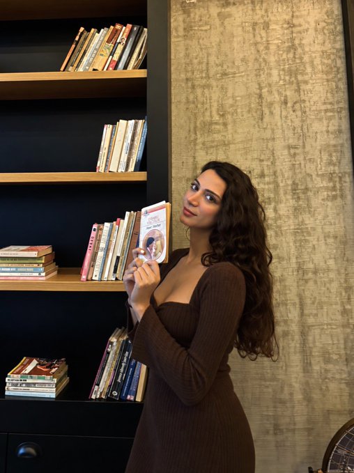 A woman with long curly dark hair wearing a brown dress stands in front of a bookshelf, holding up a small book.