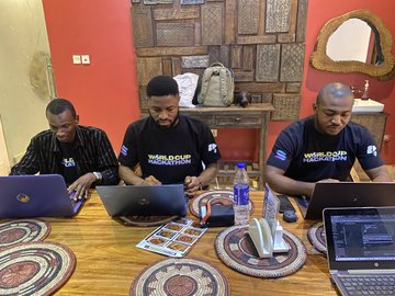 Three men in matching black 'WORLD' branded shirts working on laptops at a wooden table in a decorated indoor space.