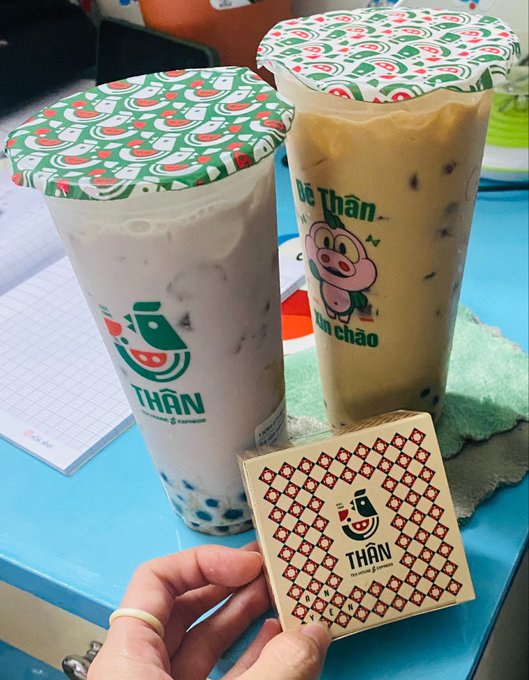 A hand holds a small brown box labeled 'THAN' and 'TEA HOUSE & COFFEE' in front of two plastic cups of iced beverages with matching patterned lids, one featuring a pig logo and 'bán chảo'.