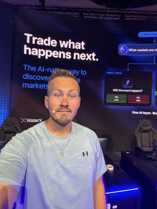 A man in an Under Armour shirt takes a selfie in front of a large display for a trading platform, featuring the text 'Trade what happens next' and a sports prediction market showing 64% Yes / 36% No.
