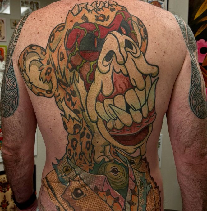 Photograph of a person's bare back featuring a large, colorful, intricate tattoo depicting a stylized skull-like face with tribal and ornamental elements.