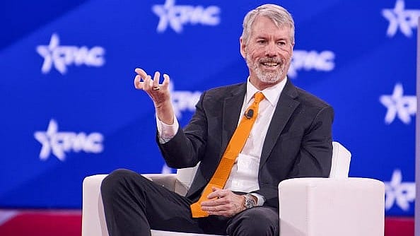 The image shows a man wearing a suit sitting in a chair while giving a speech at an event. He is wearing a yellow tie, which stands out against his formal attire. A podium can be seen next to him, indicating that he is likely addressing an audience. The backdrop features several logos and the text "RTPC".
