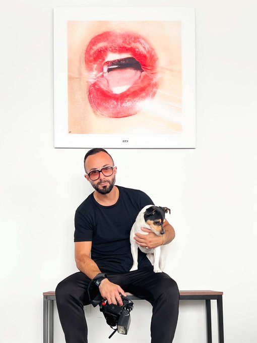 A man in a black t-shirt and glasses sitting on a bench holding a Jack Russell Terrier, posed beneath a framed artwork of glossy red lips labeled 'SEX'.