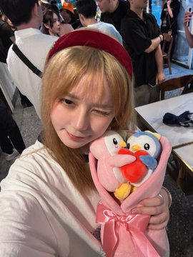 A blonde woman wearing a red beret and white top winks while holding a pink plush penguin toy at what appears to be an indoor event.