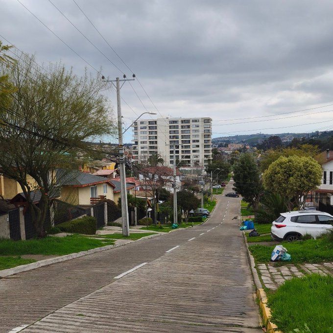 A cloudy day view down a paved residential street lined with houses, trees, and parked cars, with a tall white apartment building in the distance.