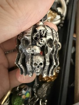 A hand holds a silver-toned metal pendant featuring a macabre design of skulls and skeletal hands, displayed in front of a dark display case with other jewelry.