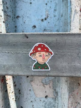 A sticker depicting a cartoon character wearing a red mushroom-cap hat with white spots and a green sweater, affixed to a weathered gray surface.