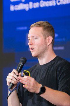 A man with short hair and stubble, wearing a black t-shirt and watch, speaks into a microphone while gesturing with his hand against a blue backdrop. Above him, partial text reads "...is calling 8 on Gnosis Chain," reinforcing the image's connection to his on-stage remarks at Pragma Lisbon about Gnosis Chain's technical goals.