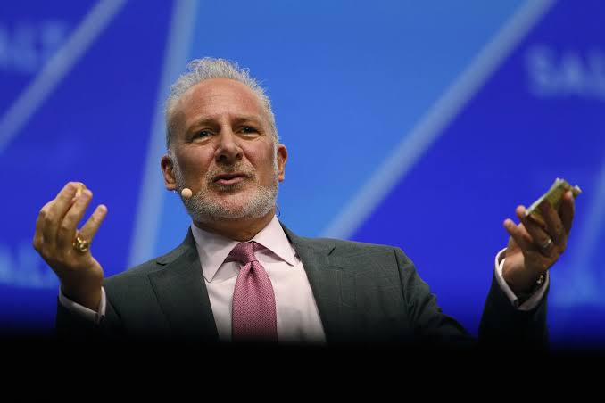 A man in a dark suit and pink tie speaking on stage with a blue background, gesturing with his hands while holding what appears to be a phone or device.