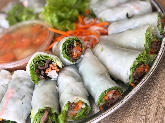 A close-up photograph of a metal platter filled with fresh Vietnamese-style rice paper rolls (gỏi cuốn) packed with greens, meat, and carrots, served alongside a bowl of dipping sauce.