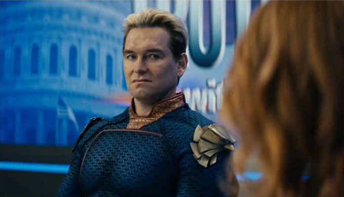A blonde man in a blue superhero costume with gold shoulder armor looks toward a red-haired person in a brightly lit room with Capitol building imagery in the background.