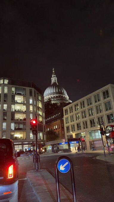 A nighttime street scene featuring St. Paul's Cathedral in London illuminated in the background, with a red traffic light and a blue directional arrow sign in the foreground.
