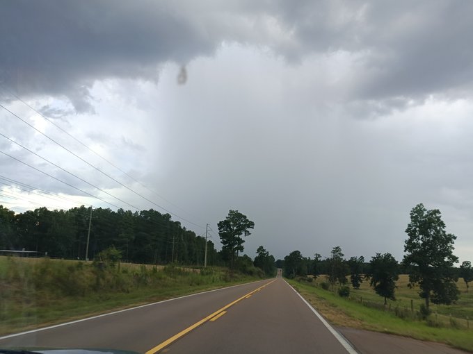 A view from a car driving down a two-lane road toward an approaching rainstorm, with dark clouds overhead and trees lining the fields.
