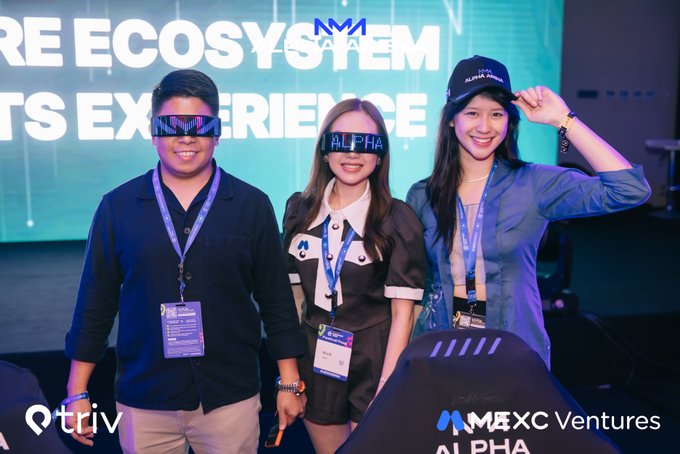 Three people pose wearing LED glasses displaying "MM" and "ALPHA" at an event with a screen reading "...RE ECOSYSTEM" and "...TS EXPERIENCE" in the background, with a "Q triv..." and "MMEXC Ventures" logo visible.