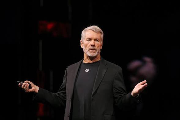 A man with gray hair and a beard, wearing a black jacket and a black t-shirt with a white logo, stands on a dark stage speaking into a headset microphone and gesturing with both hands.