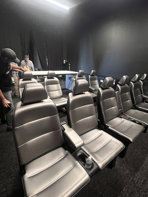 Rows of black leather theater-style recliner seats fill a dark room, where a few people stand near a curved table. The image showcases the repurposed Cybercab seats now installed in the Giga Texas theater, as referenced in the post.