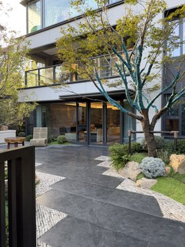 A modern multi-story residential building features a sleek dark stone walkway leading to the glass entrance, accented by a landscaped garden with a young tree, white flowering plants, and decorative river rock borders. The minimalist architecture and refined outdoor space visually echo the caption's sentiment of tangible, hard-earned accomplishment in finally having a personal studio.