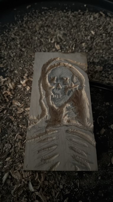 A wooden carving of a zombie-like figure with skeletal hands rests on a bed of wood shavings.