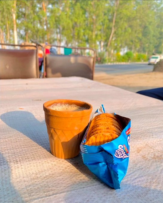 A clay cup of chai sits on a concrete ledge next to an open blue packet of Parle-G biscuits, with trees and a vehicle visible in the background.