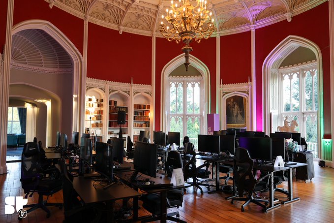 An ornate computer lab set inside a grand historic room featuring red walls, gothic arched windows, an elaborate chandelier, and rows of modern workstations with gaming chairs.