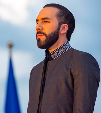 A side-profile portrait of a bearded man wearing a dark traditional-style jacket with decorative collar detailing, standing outdoors with a flag visible in the background.