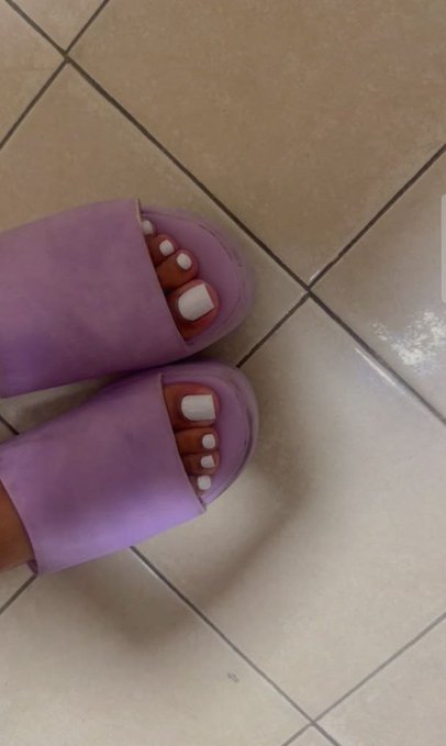 A close-up shot of feet wearing purple slide sandals with white toenail polish, standing on a beige tiled floor.