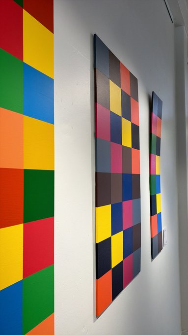 Two colorful checkered artworks hang on a white wall, one painted directly on the wall and one framed canvas with squares of various colors.