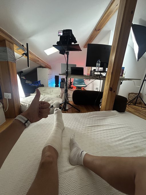 A first-person view from a bed showing feet in white socks, with a hand giving a thumbs up near a streaming/content creator setup including lights, a camera, monitors, and a microphone in a loft-style room.