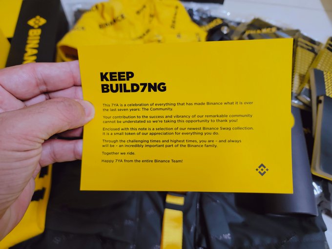 A hand holding a yellow Binance 'KEEP BUILD7NG' thank-you note celebrating Binance's 7-year anniversary, with yellow swag bags in the background.