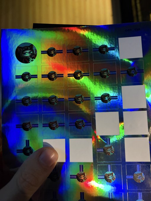 A holographic sticker sheet featuring a cat logo and multiple badge designs being held by a hand, with iridescent rainbow coloring reflecting blue, green, and orange hues.