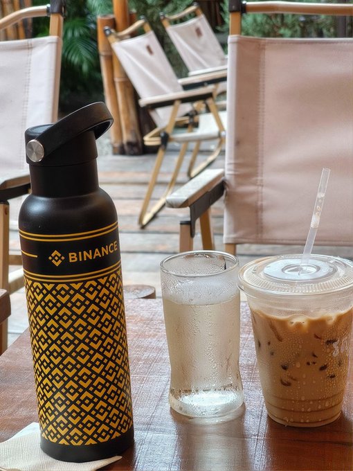 A black Binance-branded water bottle sits on an outdoor café table alongside an iced coffee drink and a glass of water. The post humorously suggests that someone "soft shilled" the $BREW token by prominently displaying the crypto sponsor's water bottle at their coffee outing.