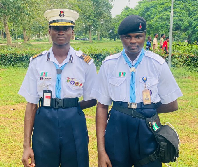 Two Nigerian Navy personnel stand side-by-side outdoors in white service uniforms, with the sailor on the left wearing a white peaked cap and the sailor on the right wearing a black beret and carrying a black bag.